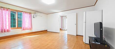 Una habitación amplia con piso de madera, un ventanal con cortinas rosadas, una puerta blanca y un televisor sobre un mueble de madera.