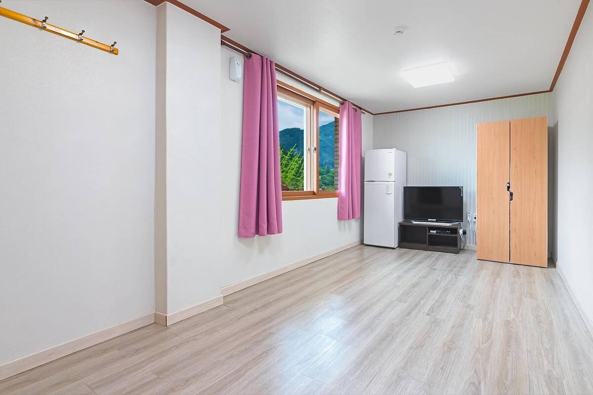 Une pièce avec un plancher en bois, un mur blanc, une armoire en bois, un réfrigérateur blanc, un téléviseur à écran plat posé sur un meuble et une fenêtre ornée de rideaux roses.