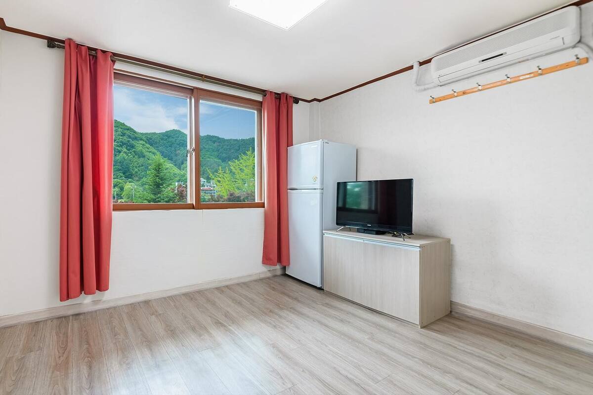 Une pièce avec un plancher en bois, un réfrigérateur blanc, un téléviseur posé sur un meuble en bois et une fenêtre ornée de rideaux rouges.