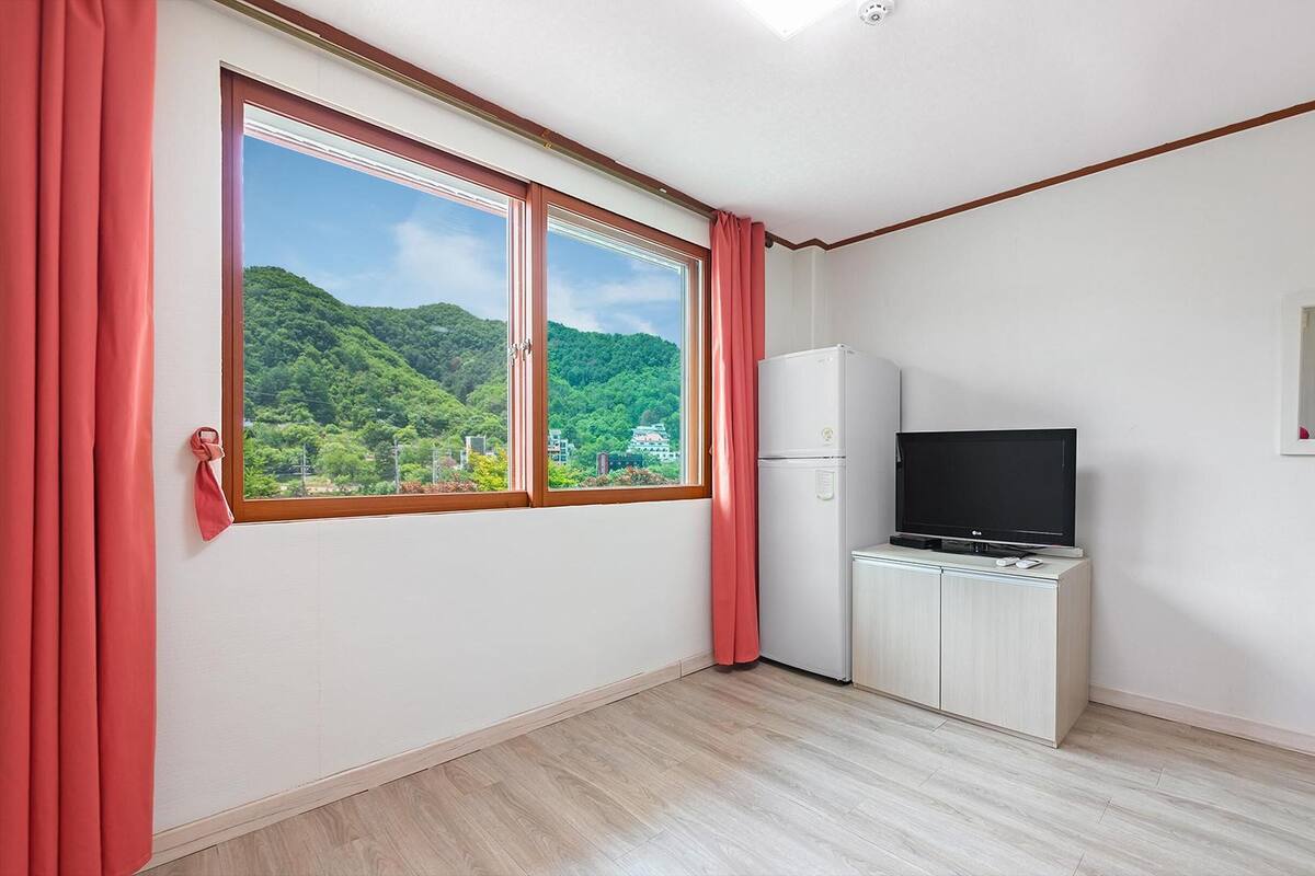 Une pièce avec une grande fenêtre, un réfrigérateur blanc, un téléviseur posé sur un meuble et un plancher en bois.