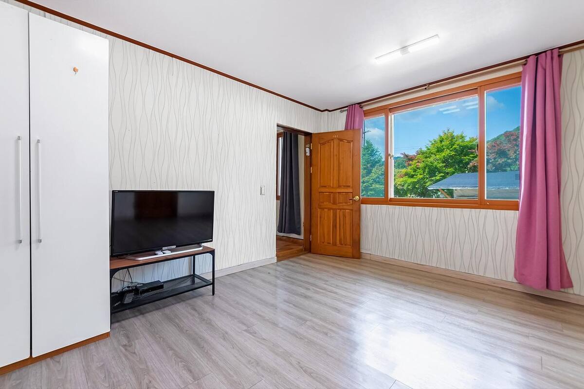 Une pièce avec un téléviseur posé sur un meuble, un plancher en bois, une armoire, une porte et une fenêtre donnant sur des arbres.