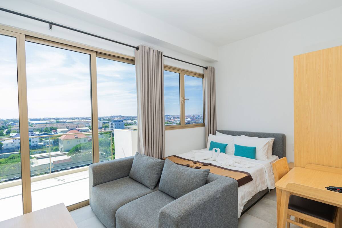 Ein modernes Hotelzimmer mit großem Fenster, einer Couch, einem Bett mit Kissen und einem Nachttisch aus Holz.