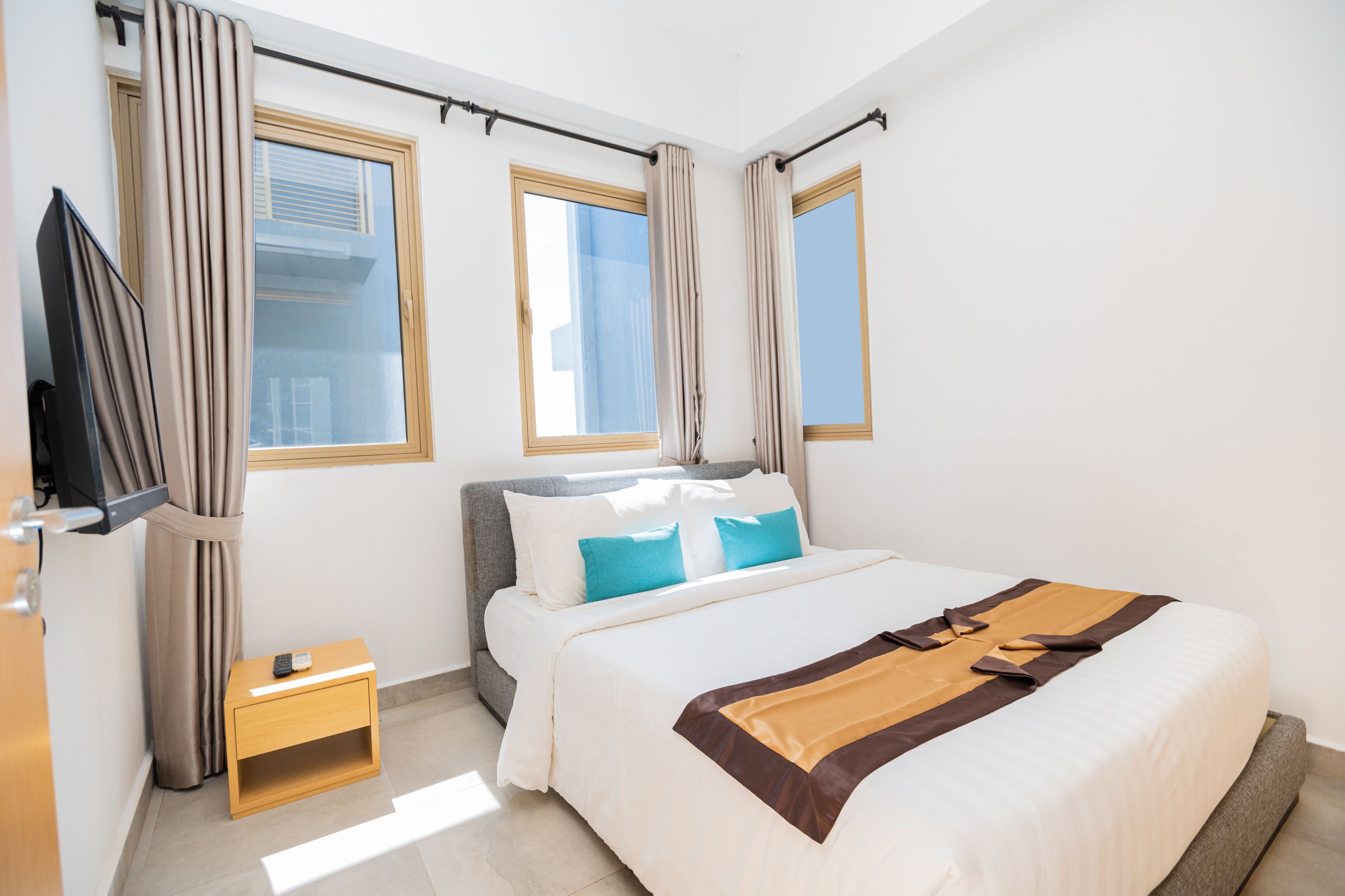 A hotel room with a bed, a TV, a small table, and two windows with curtains.