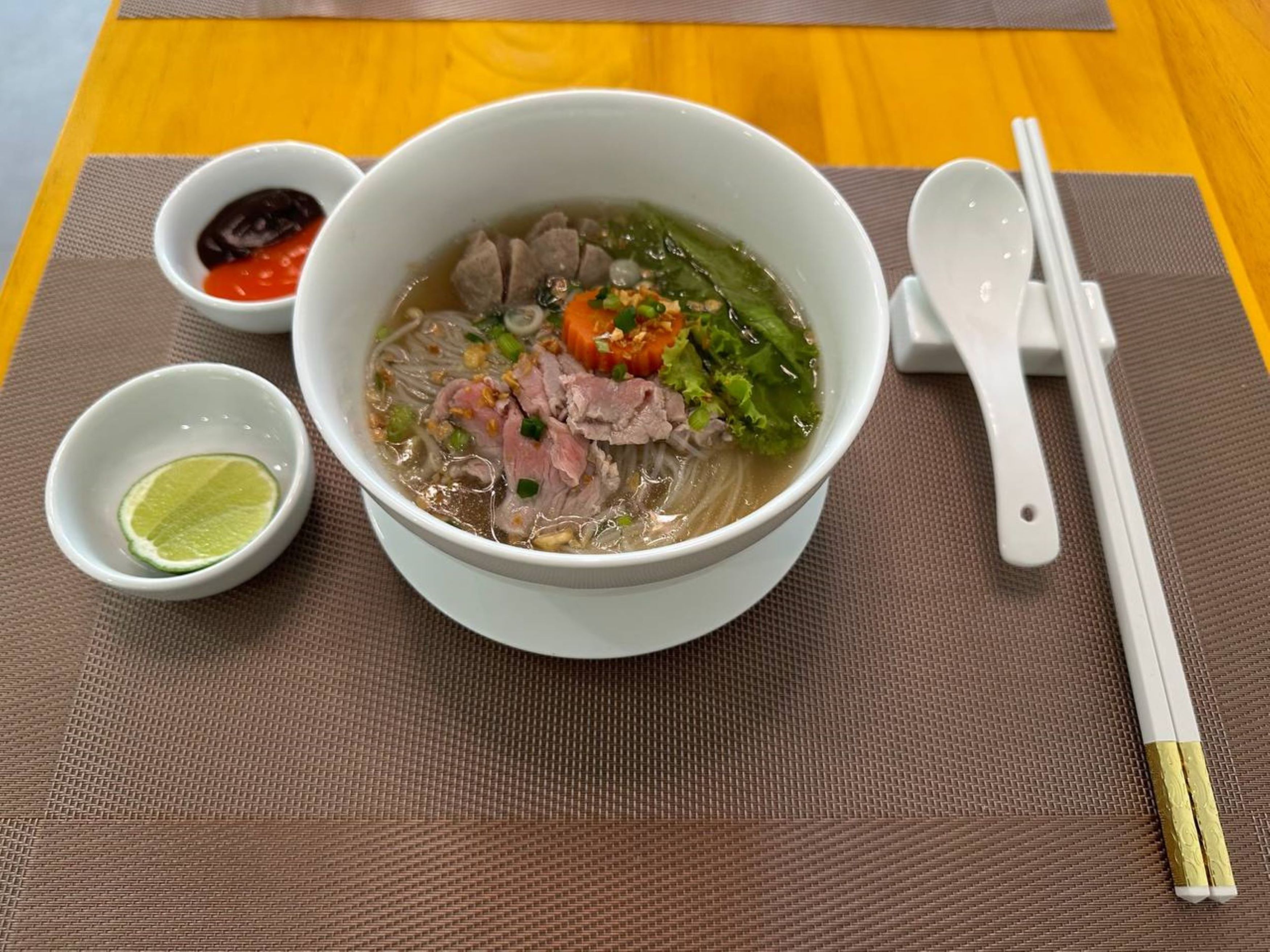 A bowl of soup with meat, vegetables, and a lime wedge on a table with a placemat, chopsticks, and a spoon.