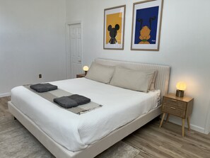 Una cama bien hecha con un edredón blanco y dos almohadas, una mesita de noche con una lámpara y dos cuadros en la pared.