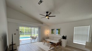 A spacious living room with a wooden floor, a ceiling fan, a white sofa, a coffee table, and a dining area with chairs and a glass door leading to a pool. - New Private Pool Mid Century Modern Home Near Disney Park (Kissimmee)