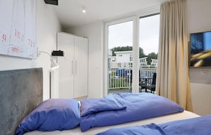 A hotel room with a bed, a desk, a chair, a TV, and a balcony with a view of buildings. - Dünenvilla Comfort (Lübeck)