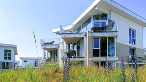 Une maison moderne à deux étages, dotée d’un balcon à l’étage supérieur, de grandes fenêtres et d’un jardin verdoyant agrémenté de fleurs sauvages.