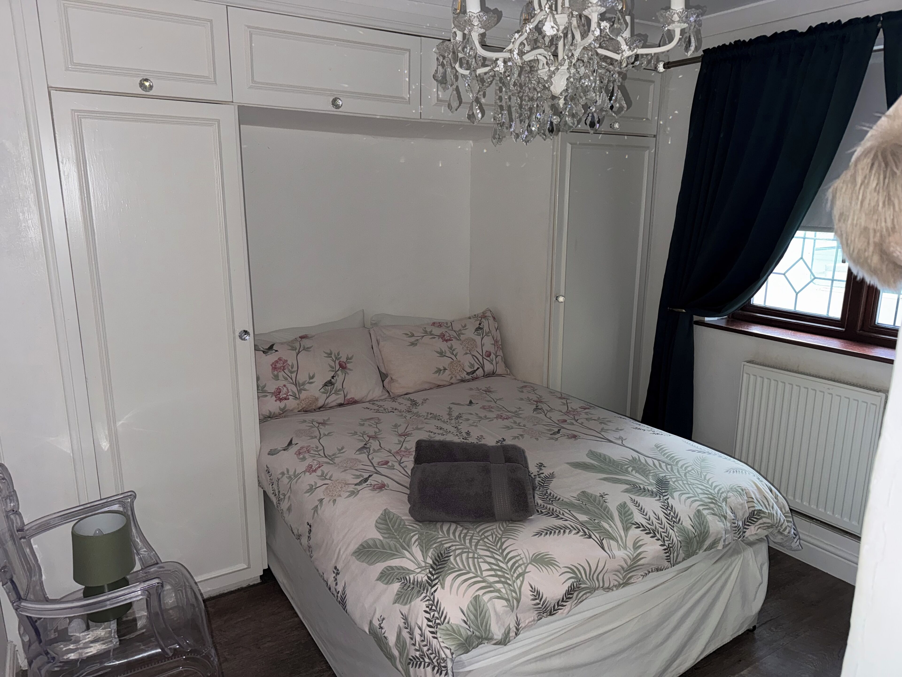 A bedroom with a bed, floral bedding, a chandelier, a window with curtains, and a radiator.