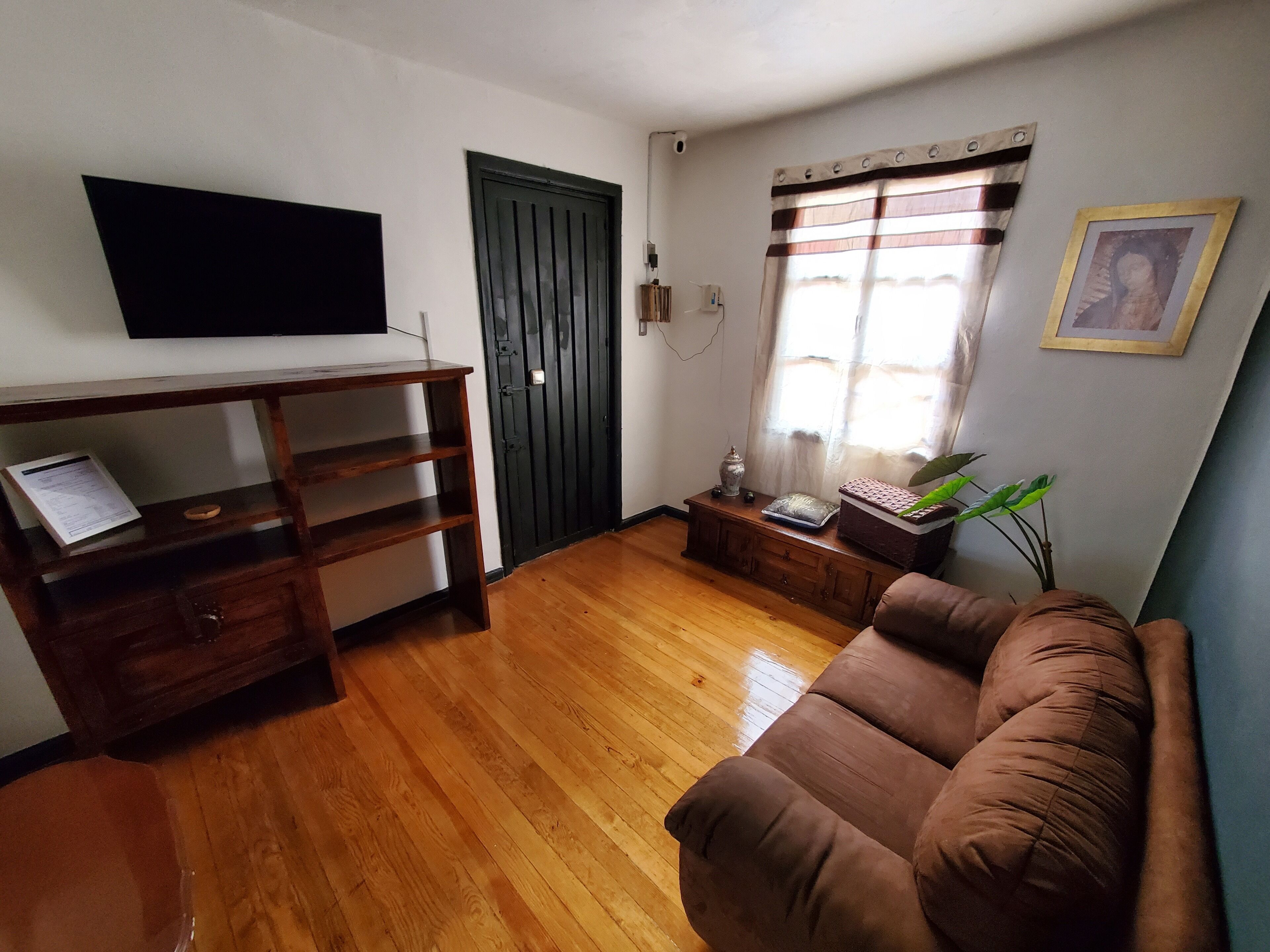 A living room with a wooden floor, a brown sofa, a TV mounted on the wall, a door, and a window with curtains.