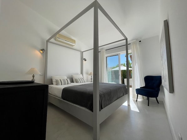 Un lit à baldaquin avec une structure blanche et une literie grise, un bureau avec une lampe et une chaise, le tout dans une chambre moderne dotée d’une grande fenêtre.