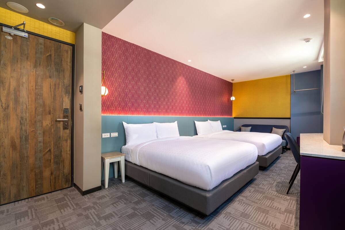 A hotel room with two beds, a wooden door, a red geometric wall, and a small table with a kettle.