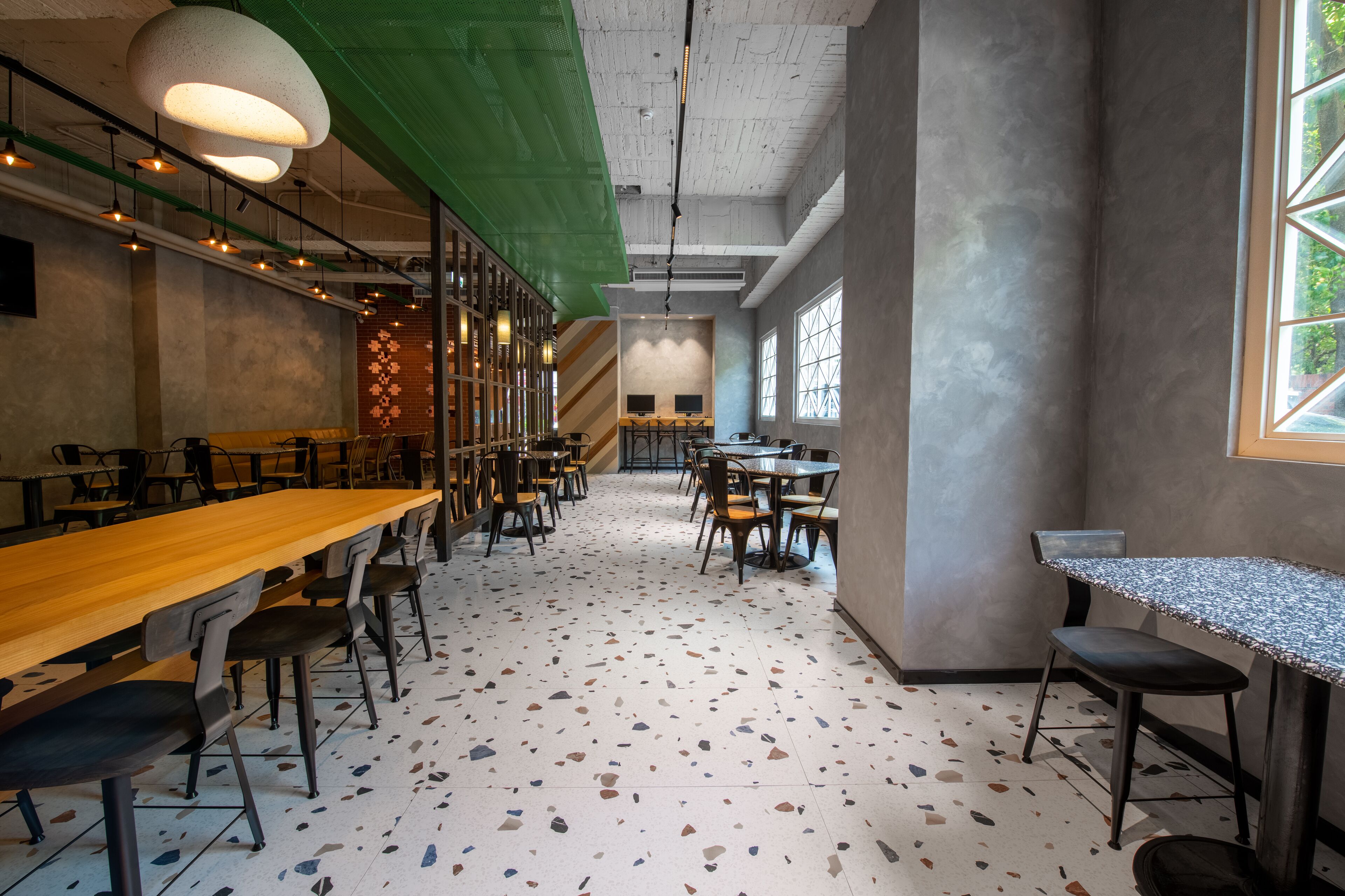 Een ruim café met lange gedeelde tafels en individuele zitplaatsen, grote ramen en een terrazzovloer.
