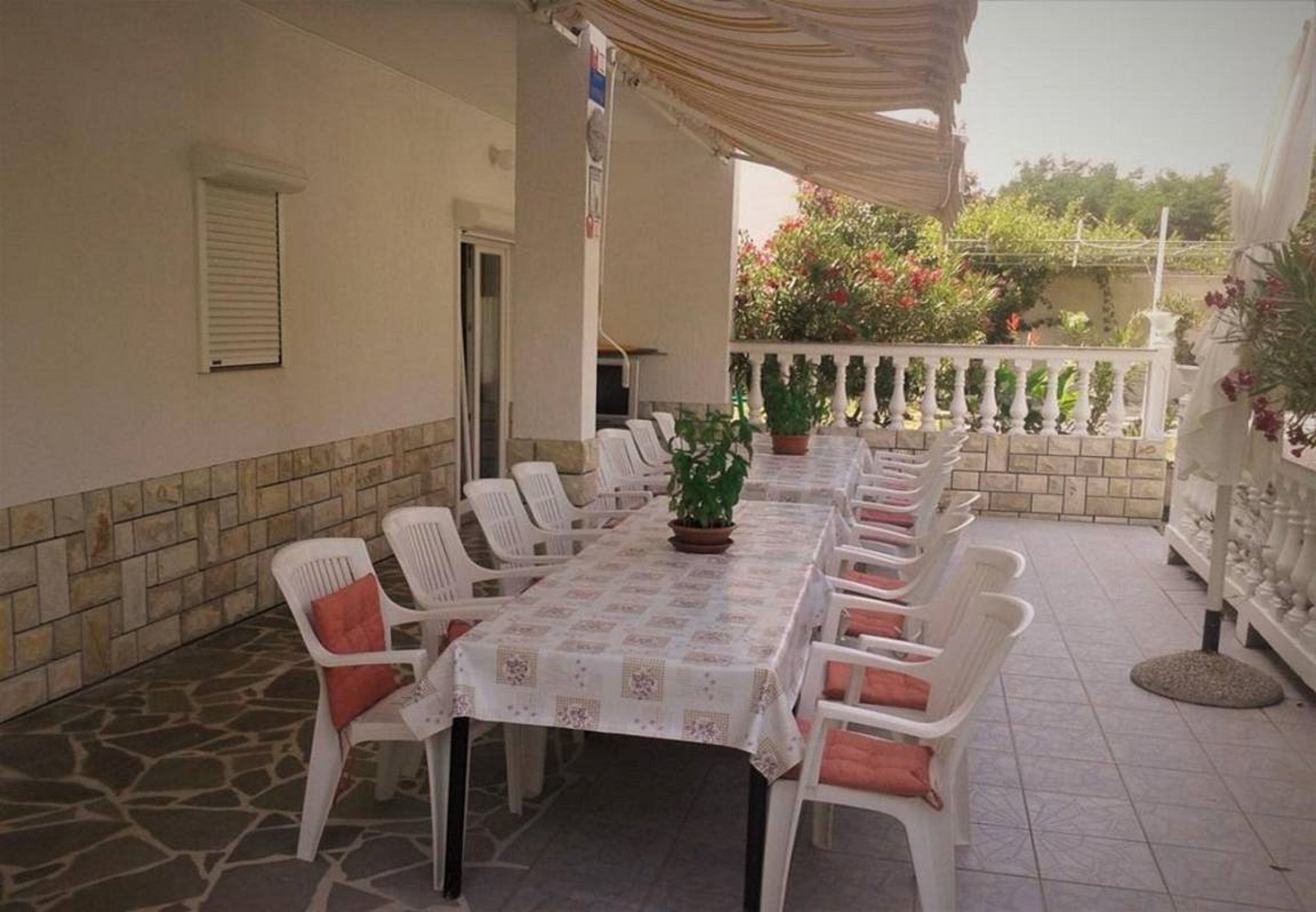 Un espace extérieur abrité, aménagé pour les repas, avec une longue table et des chaises en plastique blanc, entouré de murs en pierre et de plantes en pot.
