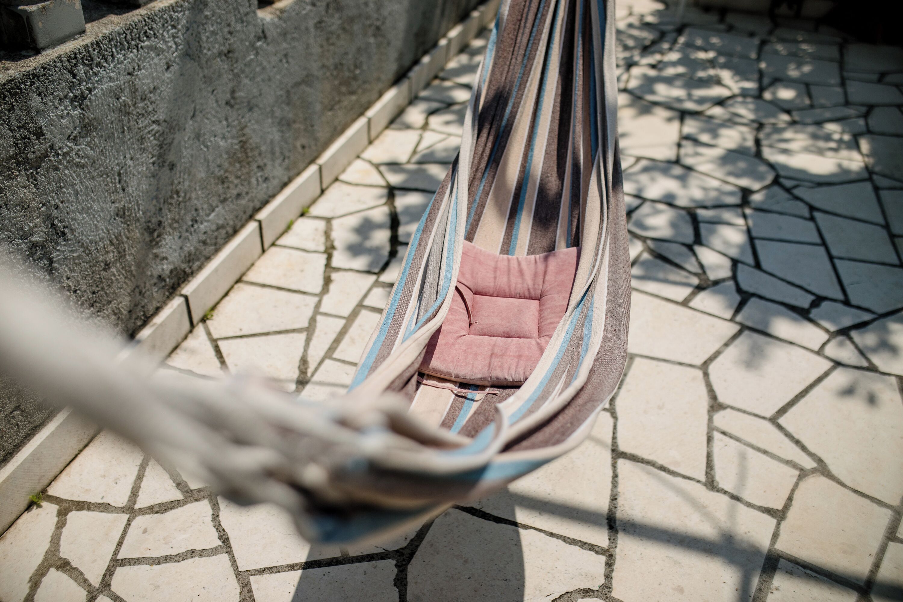 A hammock with pink cushions hanging between a stone wall and tiled floor.
