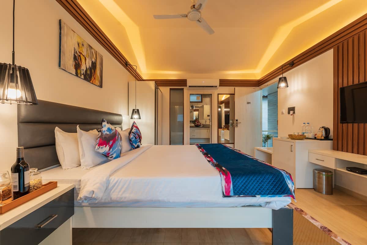 Quarto de hotel moderno com cama, mesa de cabeceira, abajur, obra de arte, ventoinha no teto e vista para o banheiro.
