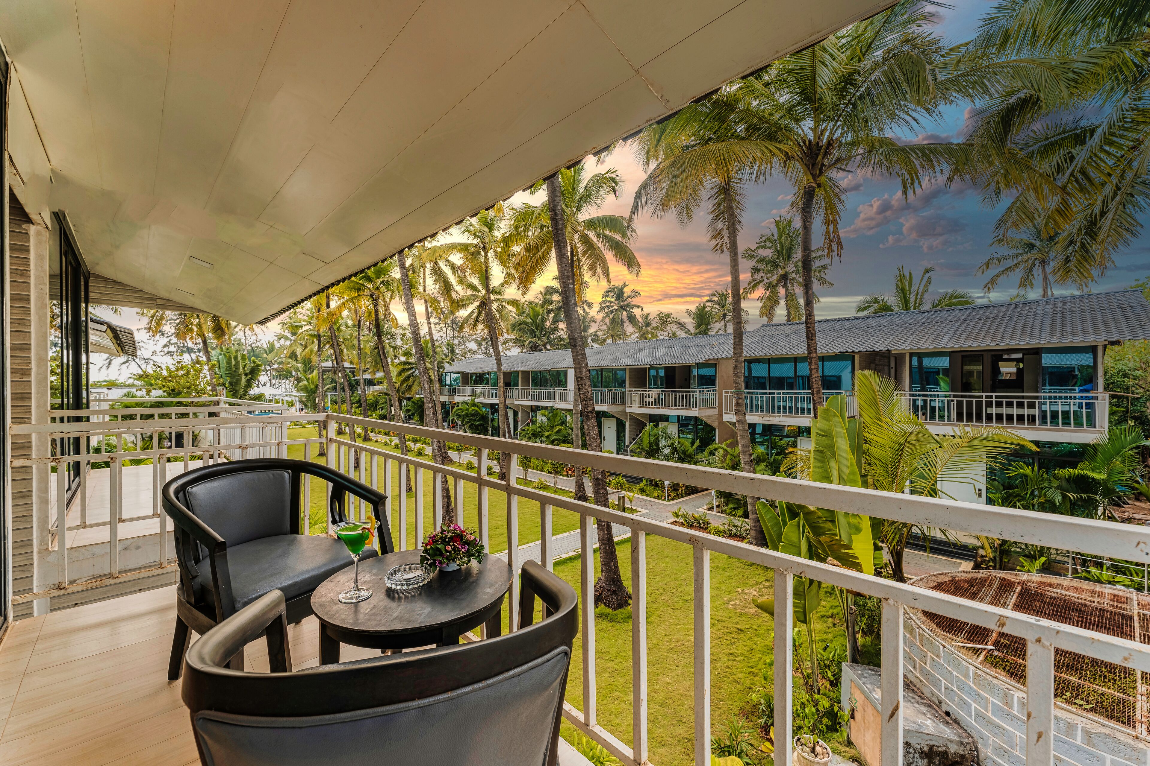 A balcony with two chairs and a table, overlooking a pool area with palm trees and a building in the background.