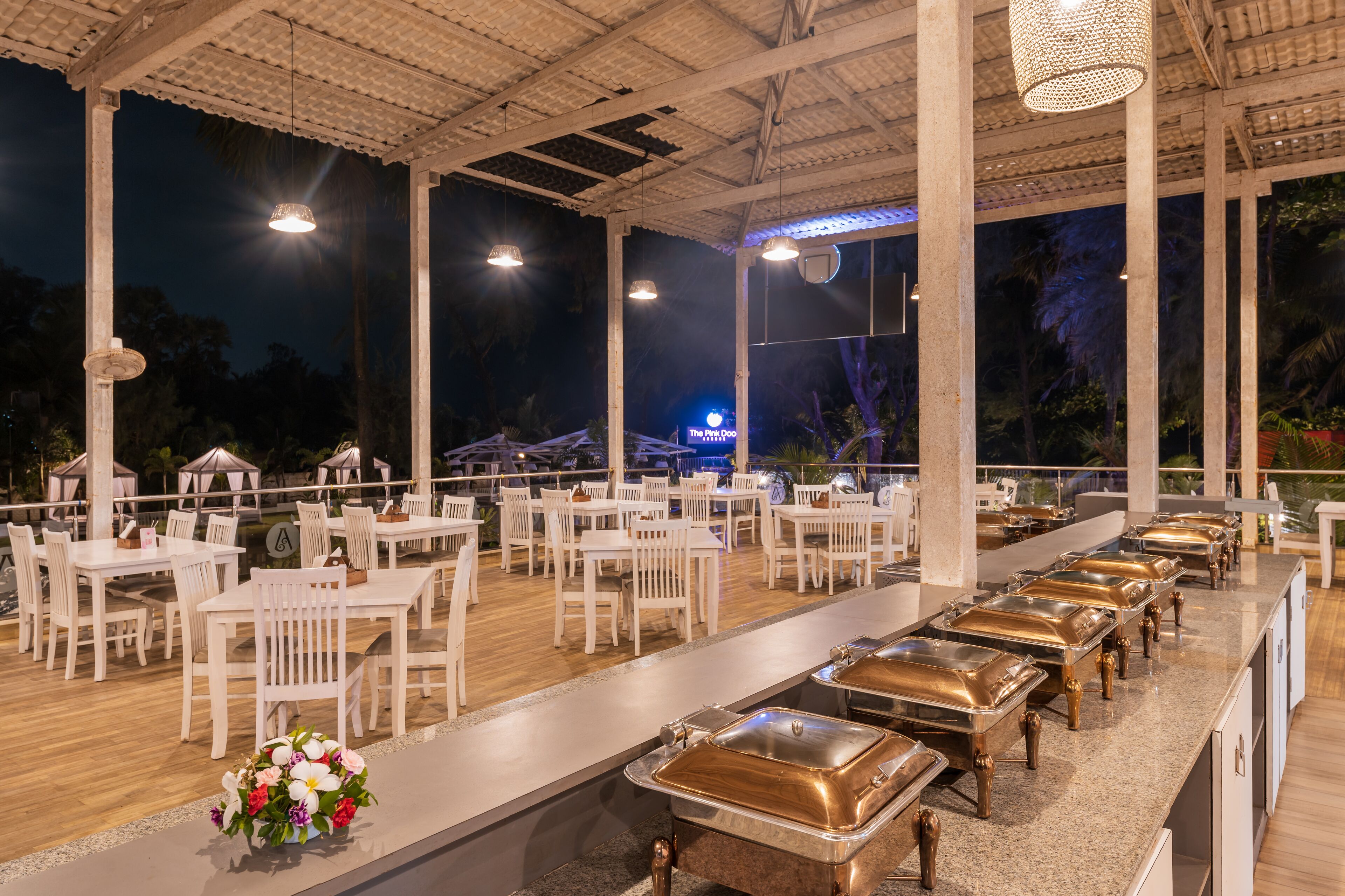 Un espace de restauration en plein air avec des tables et des chaises blanches, un buffet avec des plateaux de service en métal et un luminaire décoratif.