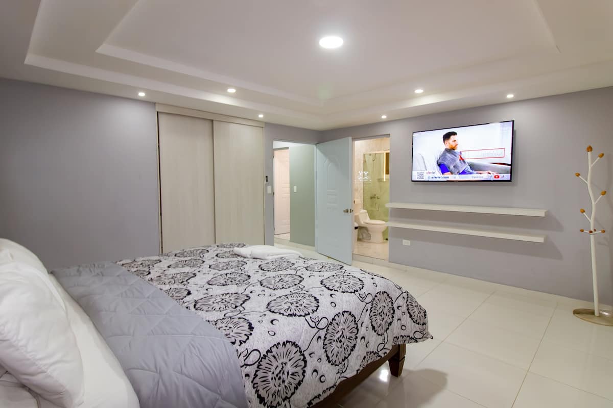 A bedroom with a bed, a TV on the wall, a wardrobe, and a bathroom visible through an open door.