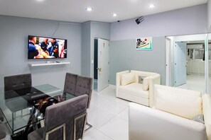 Una sala moderna con una mesa de comedor de vidrio, sillas tapizadas en gris, un sofá blanco y un televisor de pantalla plana montado en la pared.