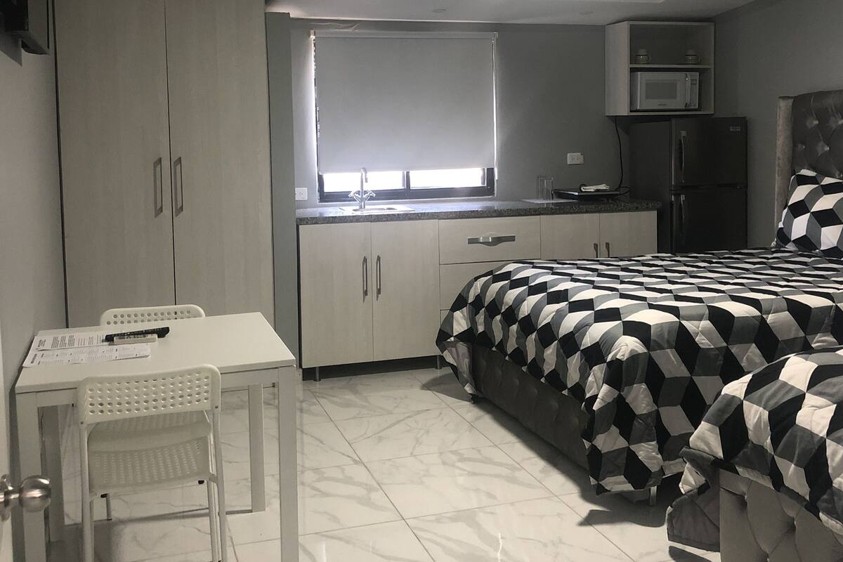 A compact bedroom with a bed, a small table, a desk, a kitchenette, and a TV mounted on the wall.