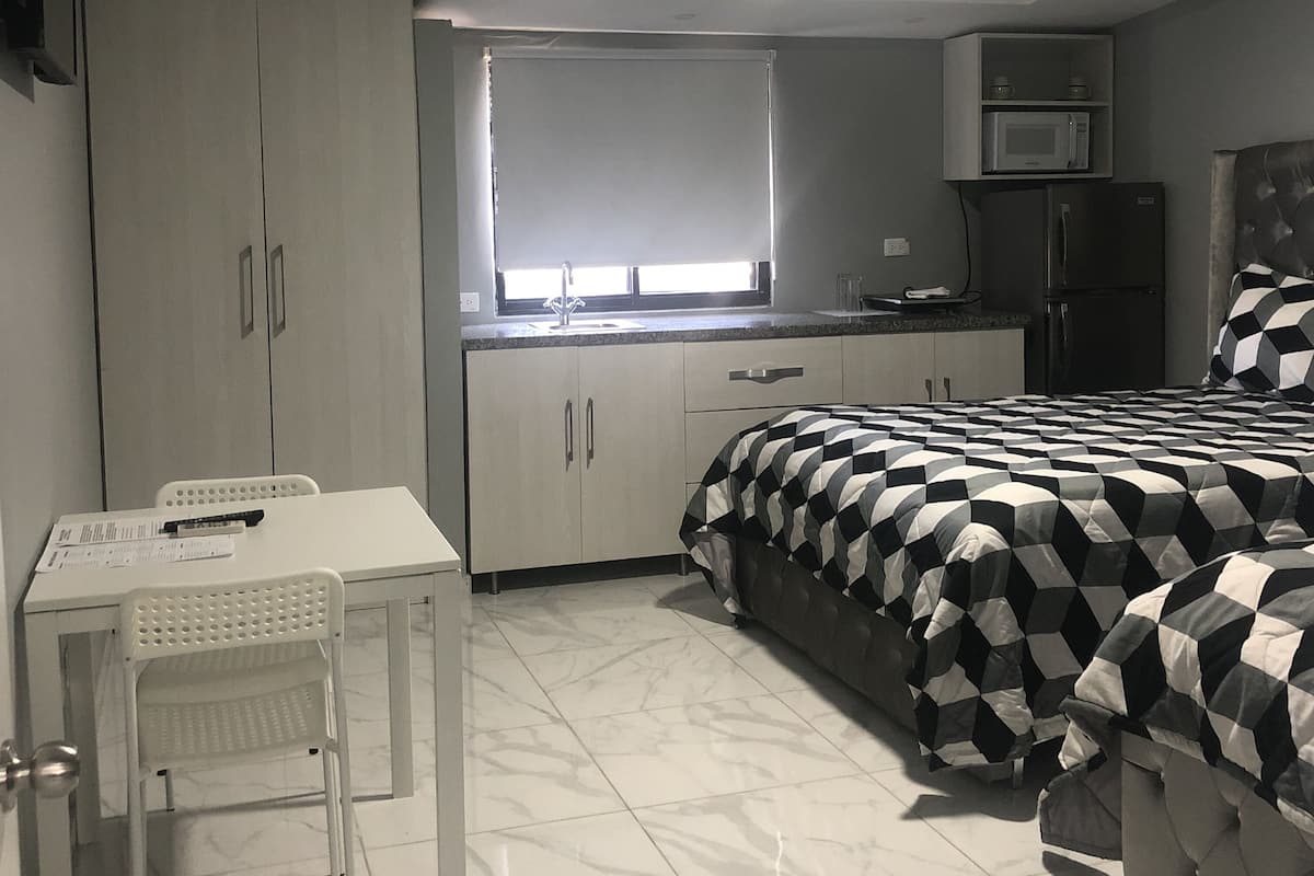 Une chambre compacte comprenant un lit, une petite table, un bureau, un coin cuisine et un téléviseur fixé au mur.