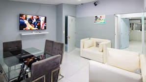 Een moderne woonkamer met een glazen eettafel, grijze en witte banken, een flatscreen televisie en een keuken die door een open deur zichtbaar is.