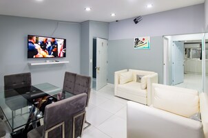 Una sala de estar moderna con una mesa de comedor de cristal, sofás grises y blancos, un televisor de pantalla plana y una cocina visible a través de una puerta abierta.