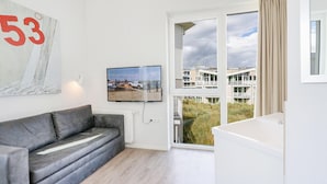 A modern living room with a grey sofa, a large painting, a television, and a view of a beach and buildings through a glass door.