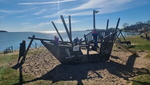Una struttura in legno a forma di barca su una spiaggia sabbiosa, con un cielo sereno e acque calme sullo sfondo.