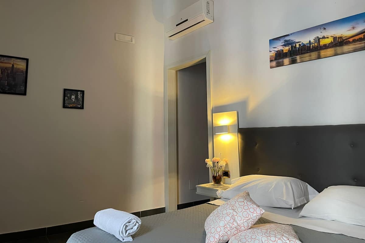 A hotel room with a bed, a nightstand, a wall-mounted air conditioner, a painting, and a framed picture.