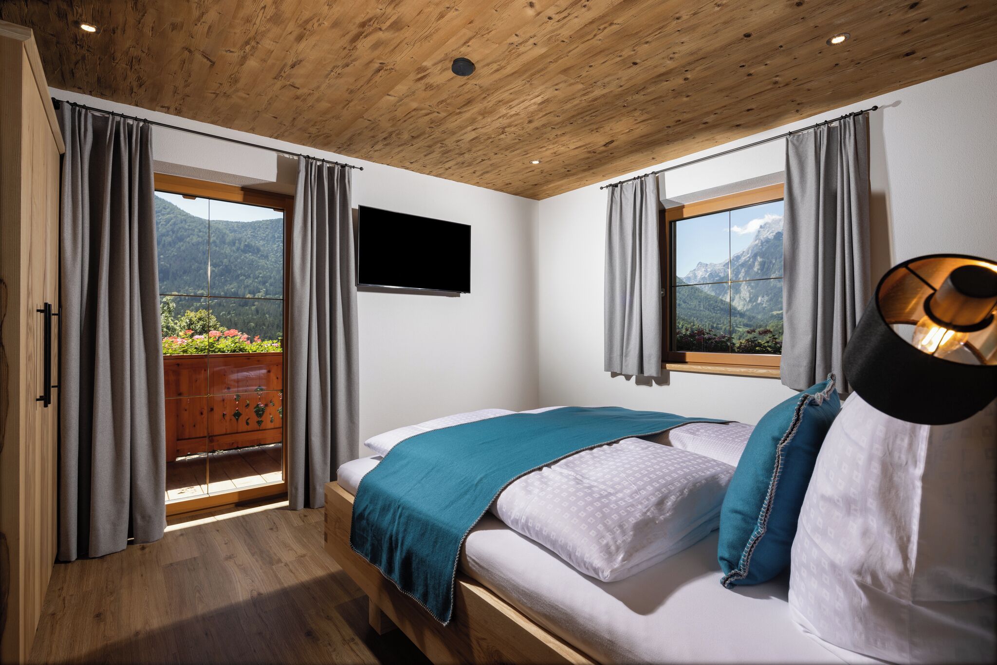 Une chambre avec un plafond en bois, un lit recouvert d’une couverture bleue, un téléviseur fixé au mur et une vue sur les montagnes par la fenêtre.