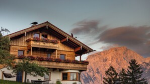 A wooden chalet with a balcony, a dining area, and a mountain backdrop.