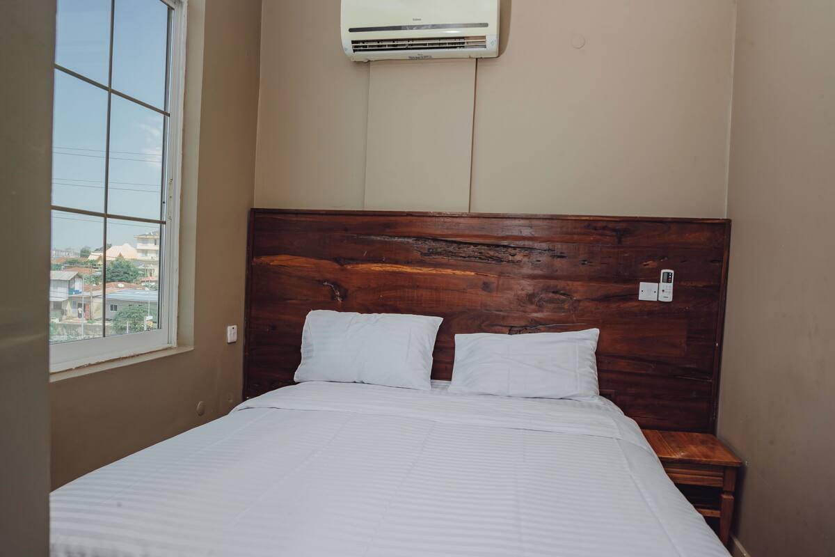 A room with a wooden headboard, a bed with white bedding, a nightstand, and a window with a view of buildings.