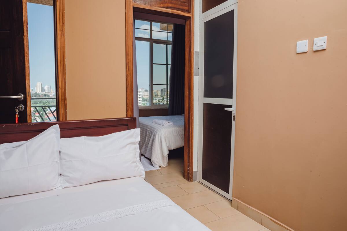 A hotel room with a bed, a window with a view of buildings, and a door leading to another room.