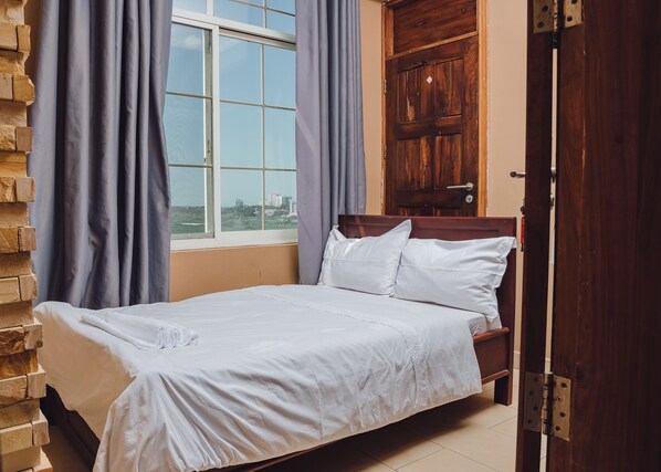 A neatly made bed with white linens, a wooden headboard, and a view of buildings through a window with curtains. - BML Highway Hotel (Dar es Salaam)