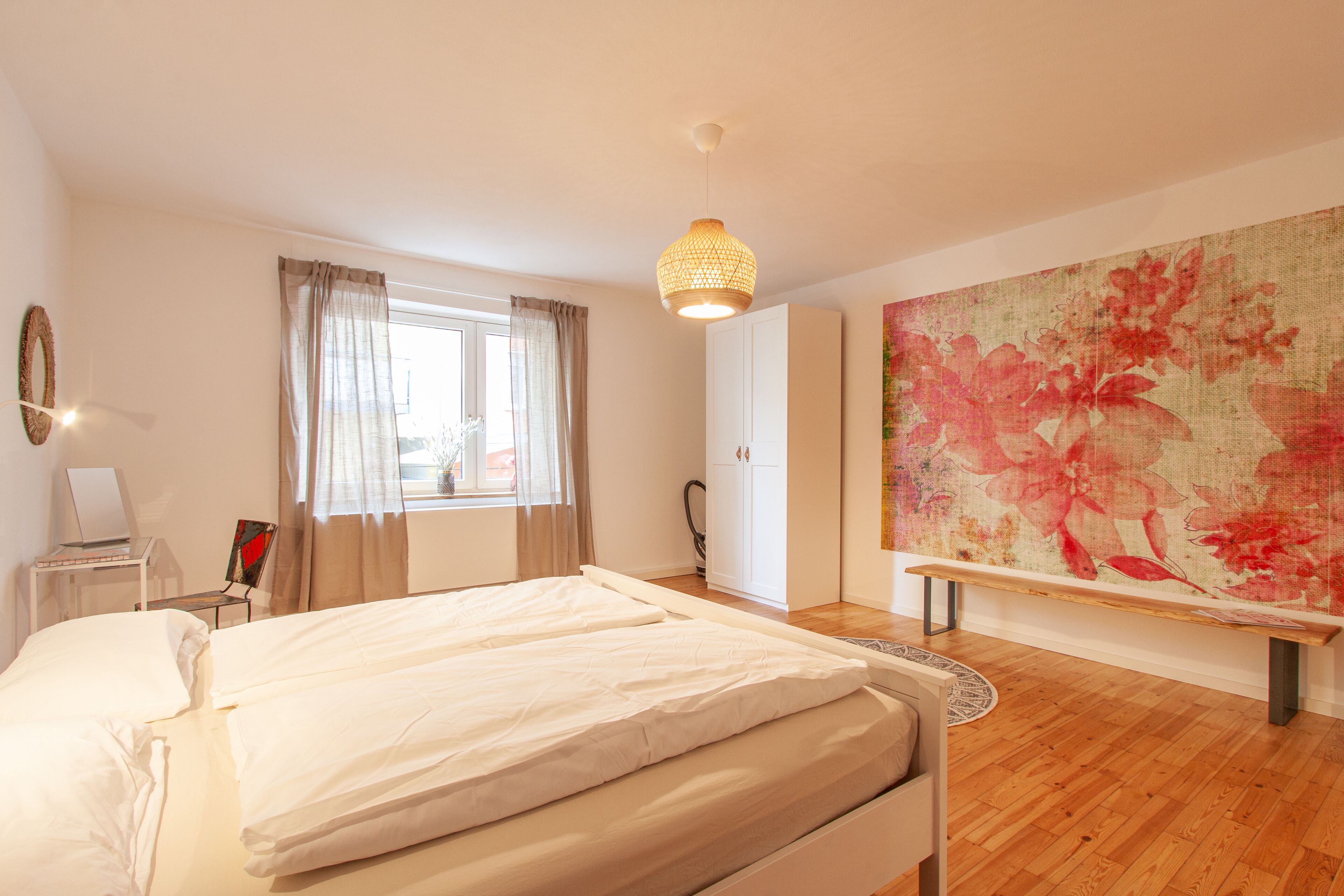 A bedroom with a bed, a desk with a laptop, a chair, a bench, and a large floral artwork on the wall.