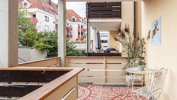 Ein Balkon mit einem gemusterten Teppich, einem kleinen Tisch mit einer Topfpflanze und Metallstühlen.