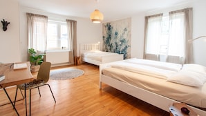 Ein Schlafzimmer mit Holzboden, zwei Betten, einem Schreibtisch, einem Sessel, einer Topfpflanze und einem großen Fenster mit Vorhängen.