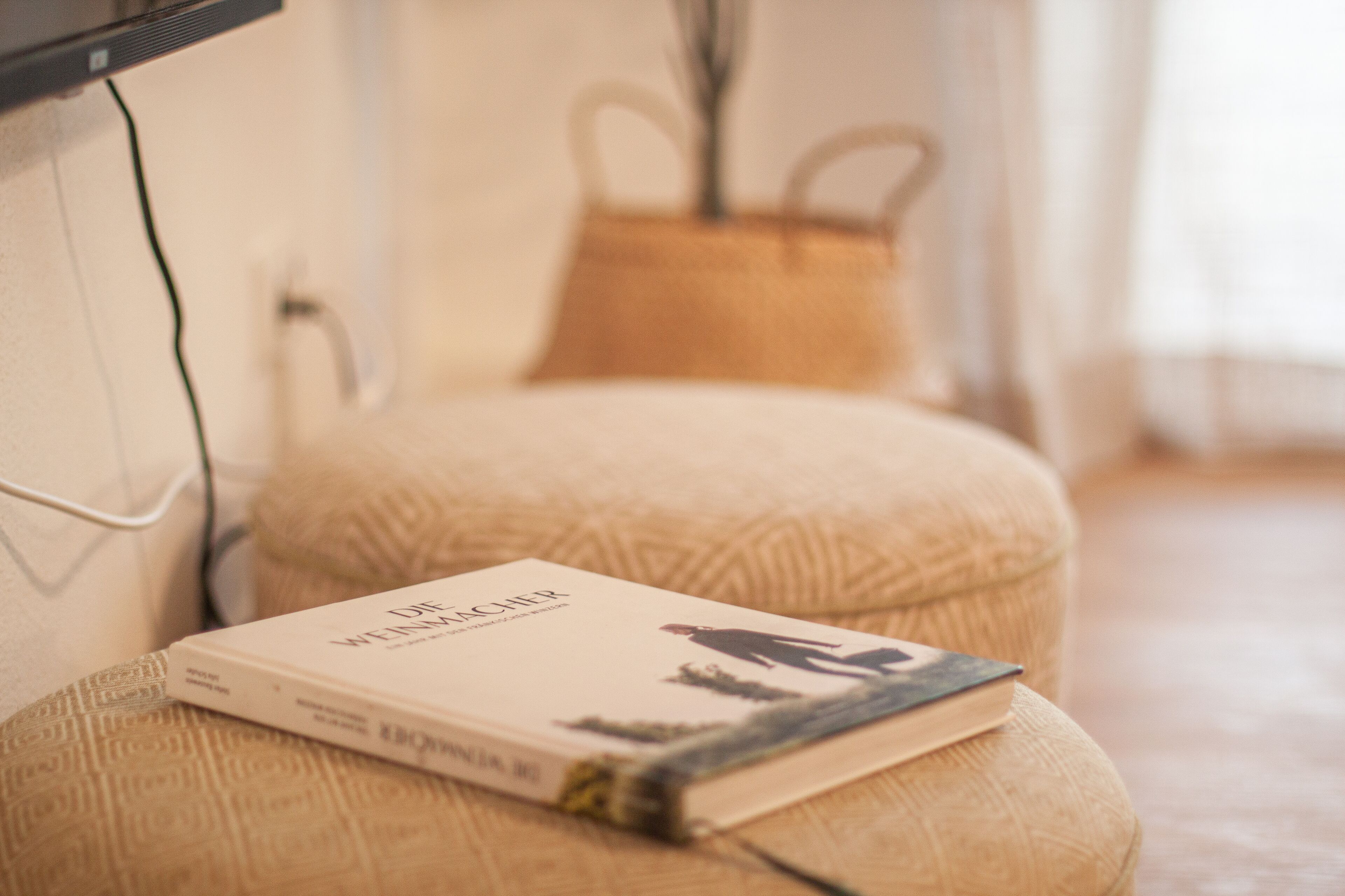 Ein Buch mit dem Titel 'Die Weichter' auf einem beigen Pouf in einem Zimmer mit Holzboden und einem gewebten Korb.