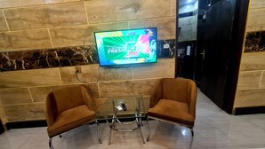 A lobby with a TV mounted on the wall displaying an advertisement. There are two brown armchairs and a small table with a remote control on it. - Smart Apartments & Hotel (Lahore)