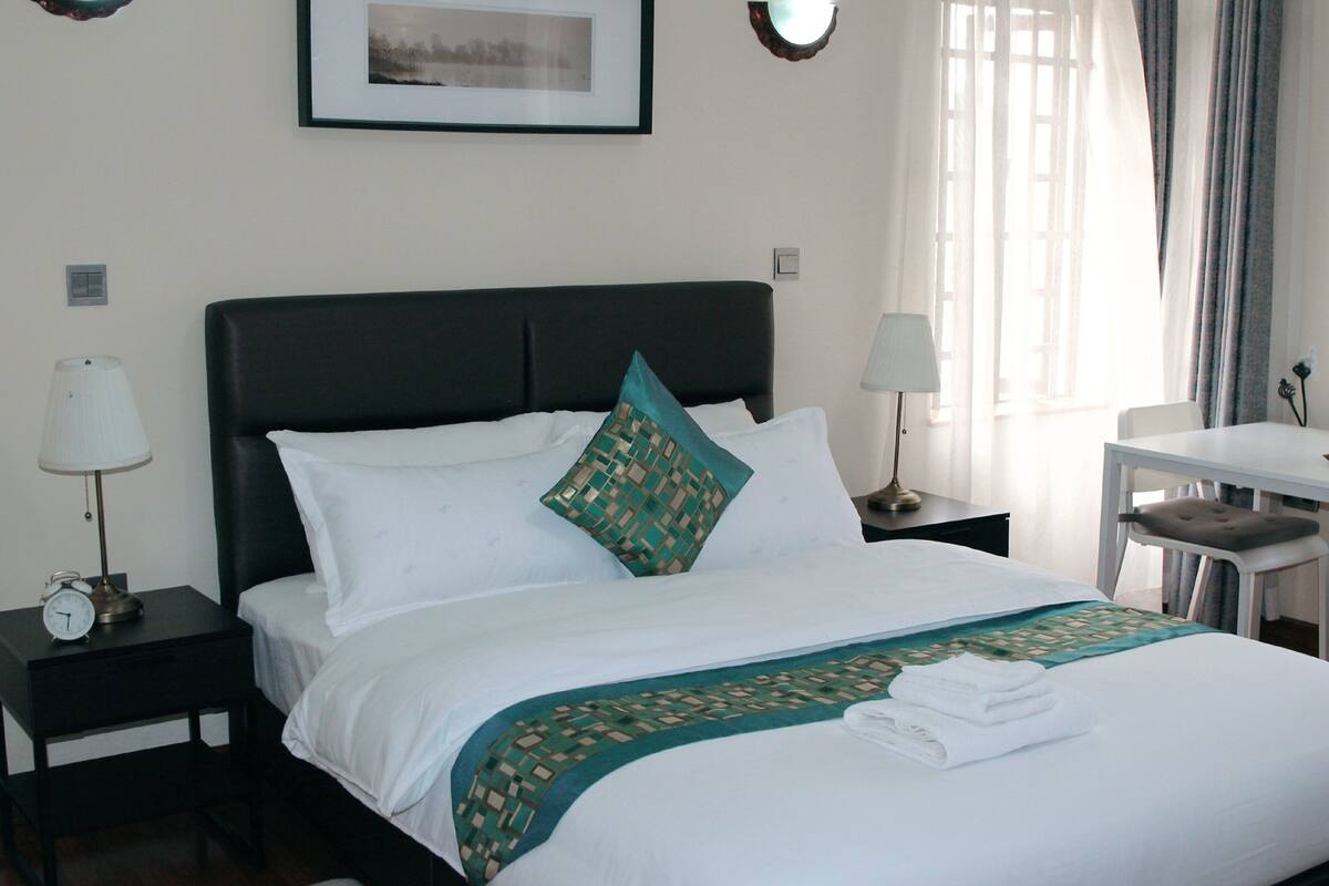 A neatly made bed with white linens and a teal patterned throw, a bedside table with a lamp, and a framed picture on the wall.