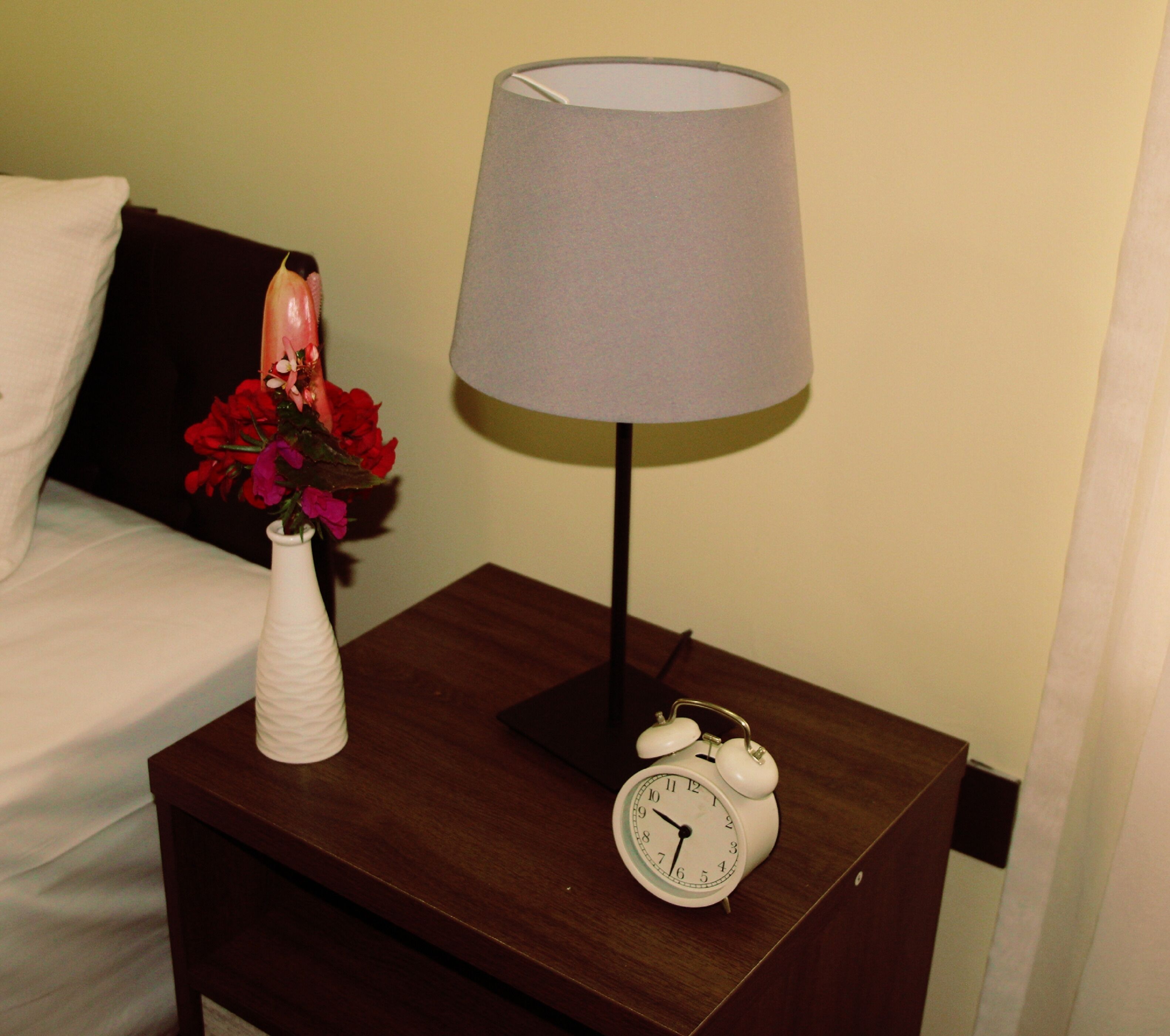 A wooden nightstand with a white alarm clock, a white vase with red flowers, and a grey lampshade.