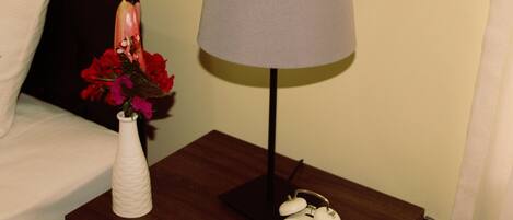 A wooden nightstand with a white alarm clock, a white vase with red flowers, and a grey lampshade.