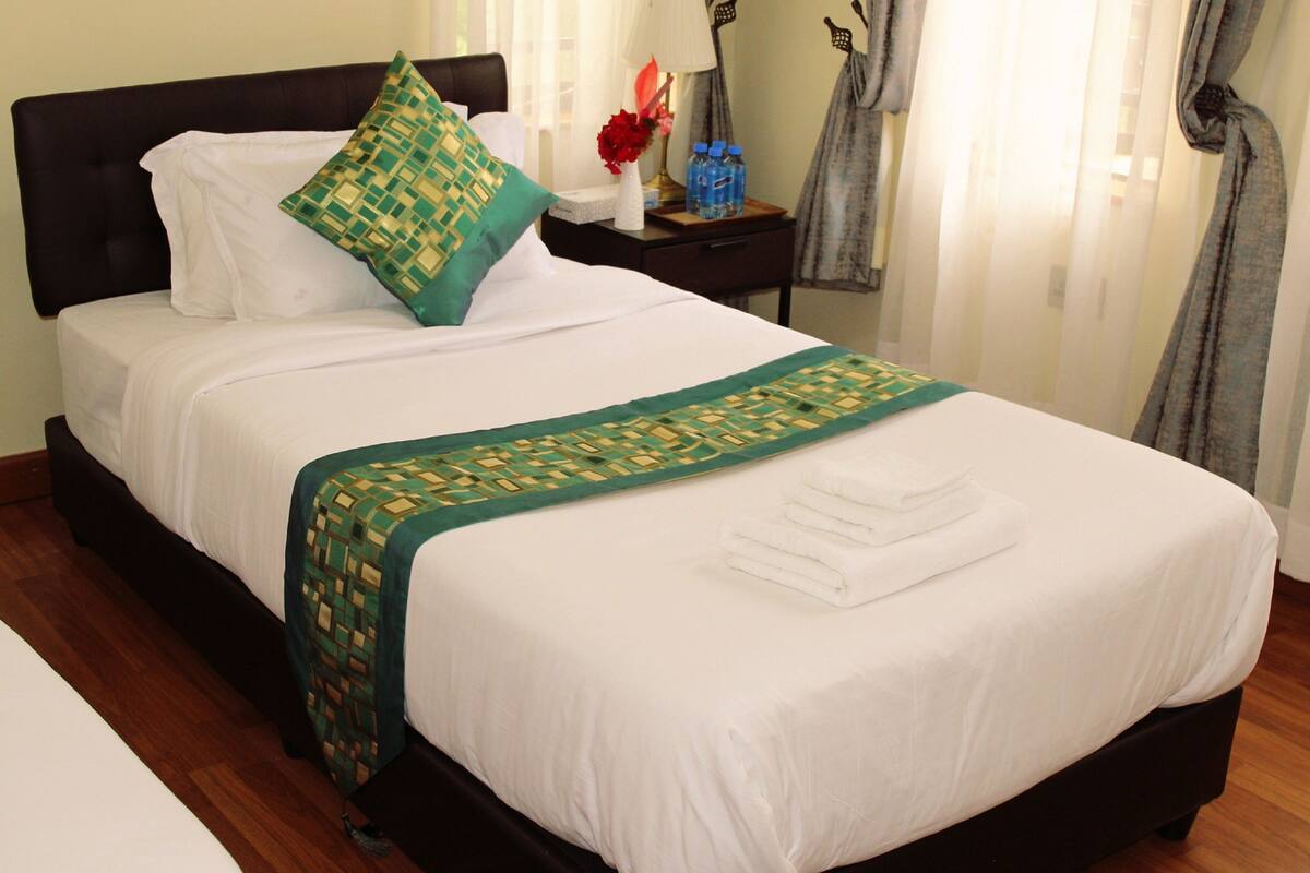 A neatly made bed with white linens, a green patterned throw, and a white towel folded at the foot of the bed.