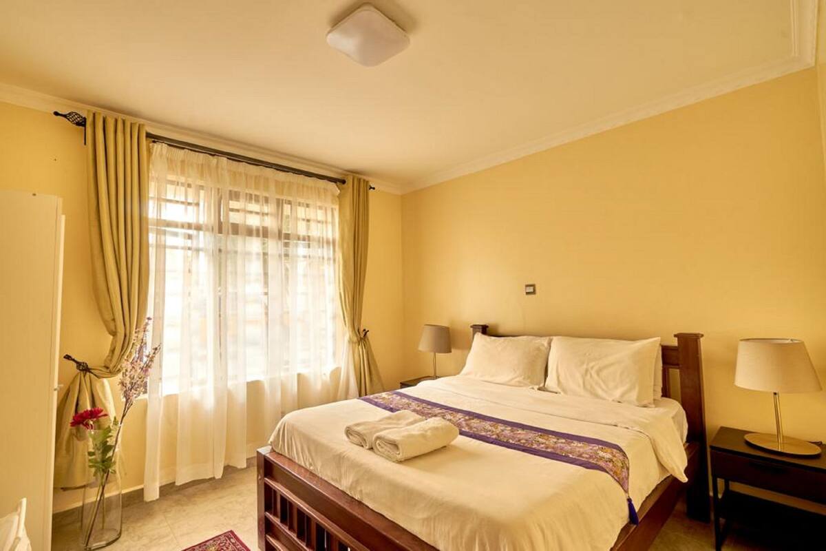 A hotel room with a bed, bedside lamps, a window with curtains, and a small table with a lamp.