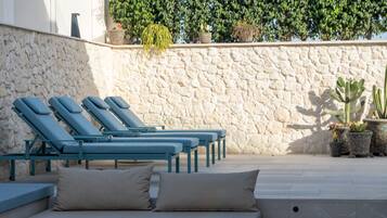 A row of blue lounge chairs and a beige sofa with cushions, arranged on a stone patio with a white wall and potted plants in the background.