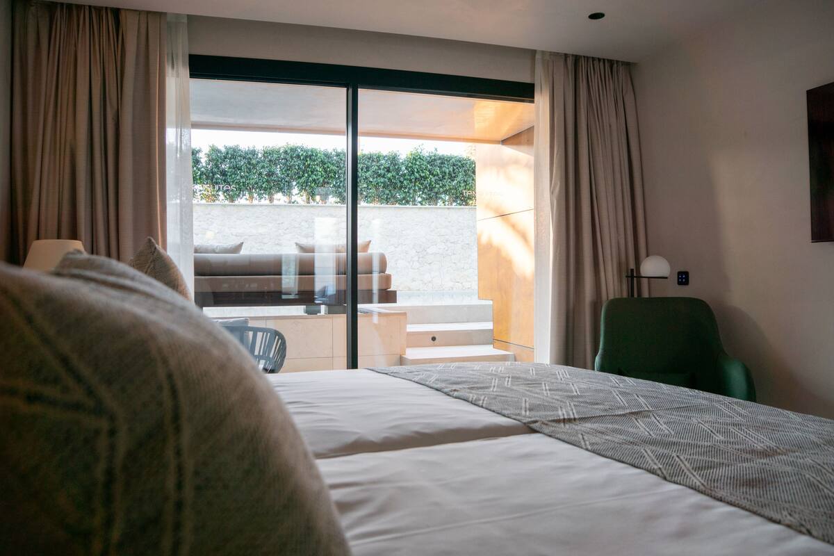 A hotel room with a bed, a green chair, a desk, and a view of a garden through a sliding door.