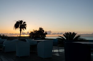 Una terraza con sillas y una palmera, con vista a una puesta de sol sobre el océano.