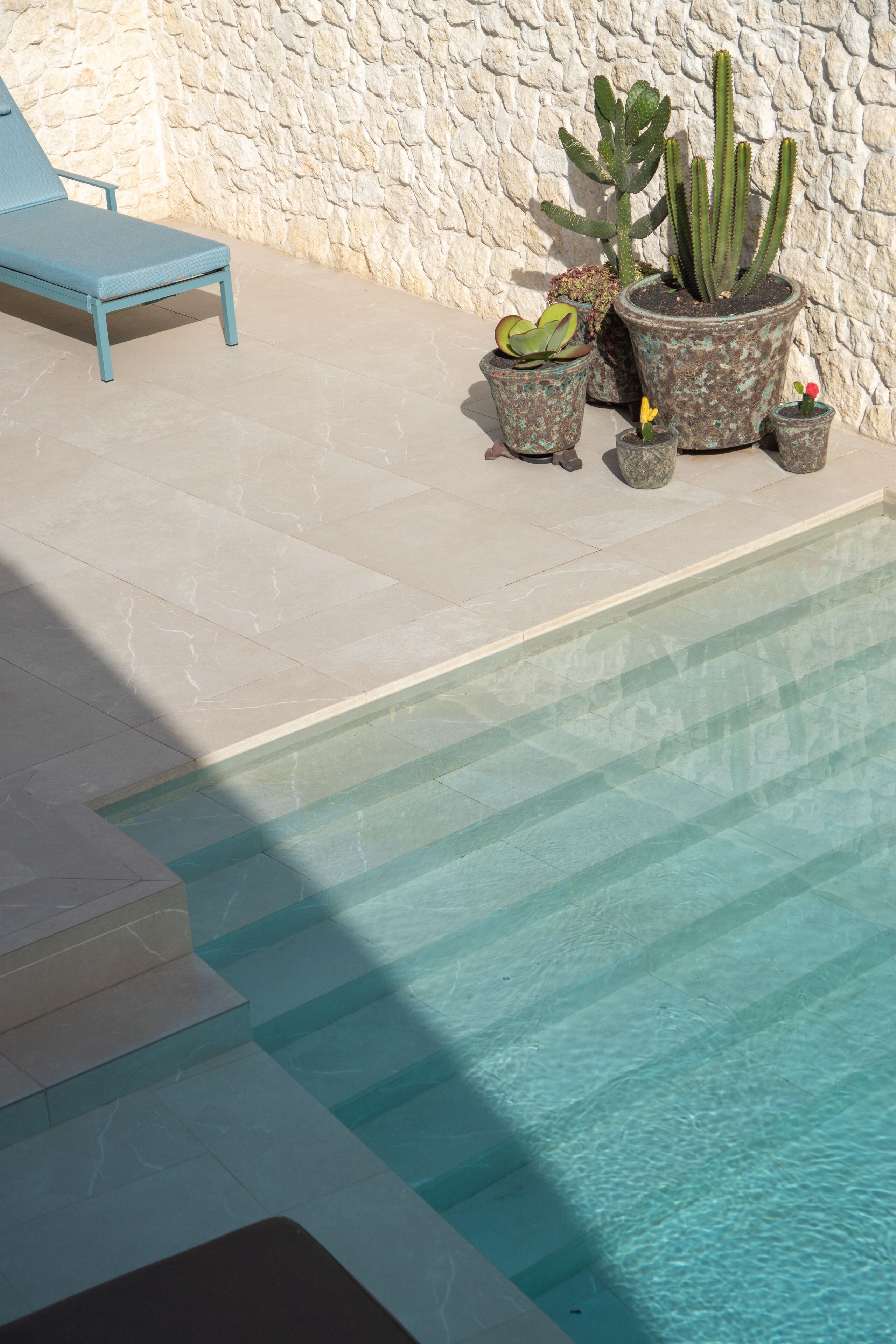 Zona de piscina con una tumbona, pared de piedra y cactus en macetas.